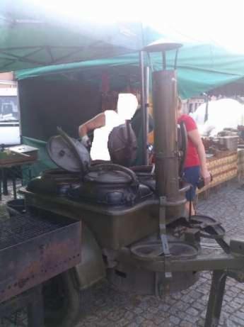 Polowa kuchnia wojskowa z otwartą pokrywą, przygotowująca posiłek pod zielonym namiotem na brukowanym placu.