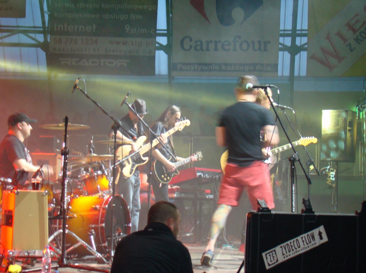 Zespół muzyczny grający na scenie podczas wydarzenia plenerowego, widoczny perkusista, gitarzyści i klawiszowiec, w tle reklama Carrefour.