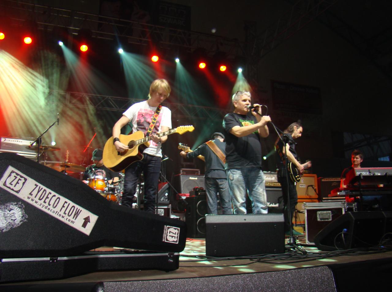 Zespół muzyczny Zydeco Flow występuje na scenie w oświetleniu reflektorów; wokalista śpiewa do mikrofonu obok gitarzysty grającego na akustycznej gitarze, a na pierwszym planie widoczny futerał...