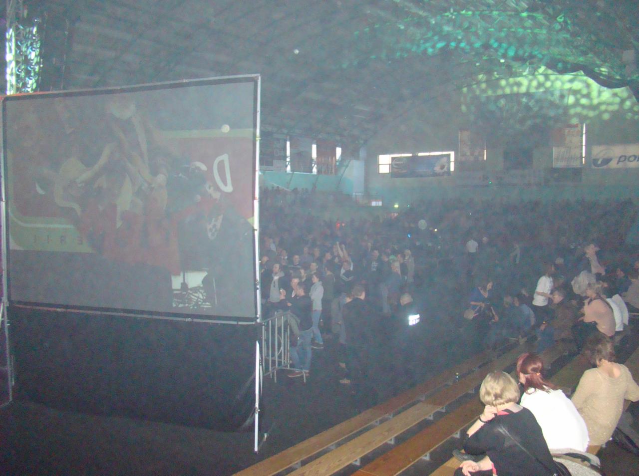 Duża sala widowiskowa wypełniona publicznością, ekran projekcyjny z widocznym fragmentem obrazu po lewej stronie, oświetlenie punktowe na suficie, rzędy ławek dla widzów na pierwszym planie.
