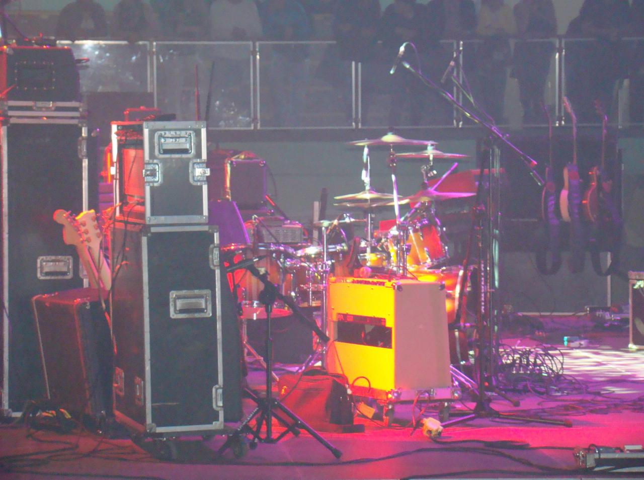 Scena przed koncertem: perkusja, gitary wiszące na stojaku, skrzynie transportowe, mikrofony i kable, oświetlone fioletowym i żółtym światłem.
