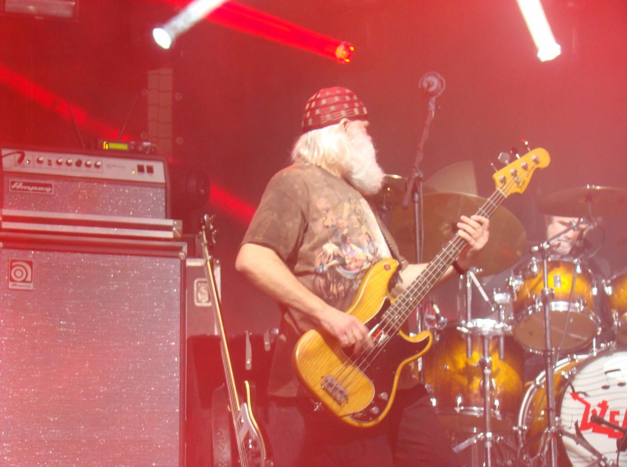 Basista z długą białą brodą w czapce gra na basie na scenie z bębnami i wzmacniaczami, oświetlony czerwonym światłem.