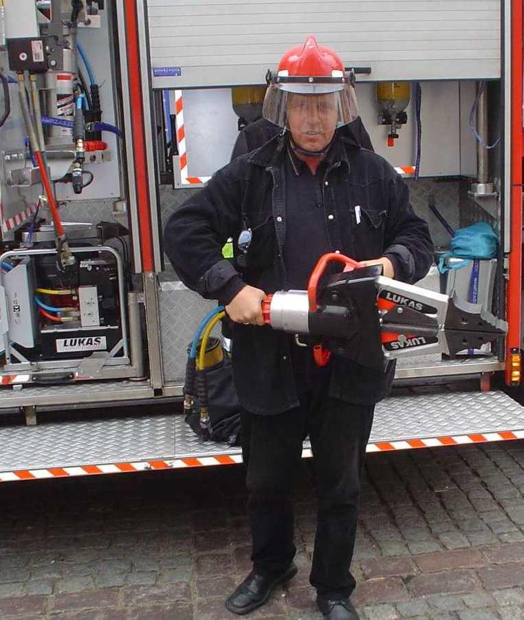 Strażak w czerwonym hełmie i osłonie twarzy, trzymający narzędzie hydrauliczne LUKAS przed wozem strażackim.
