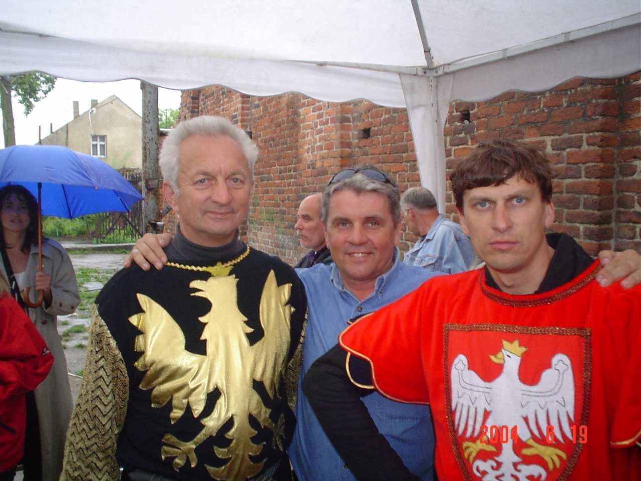 Trzech mężczyzn w kostiumach z motywem orła na tle ceglanej ściany i białego namiotu, jeden z parasolem w tle.