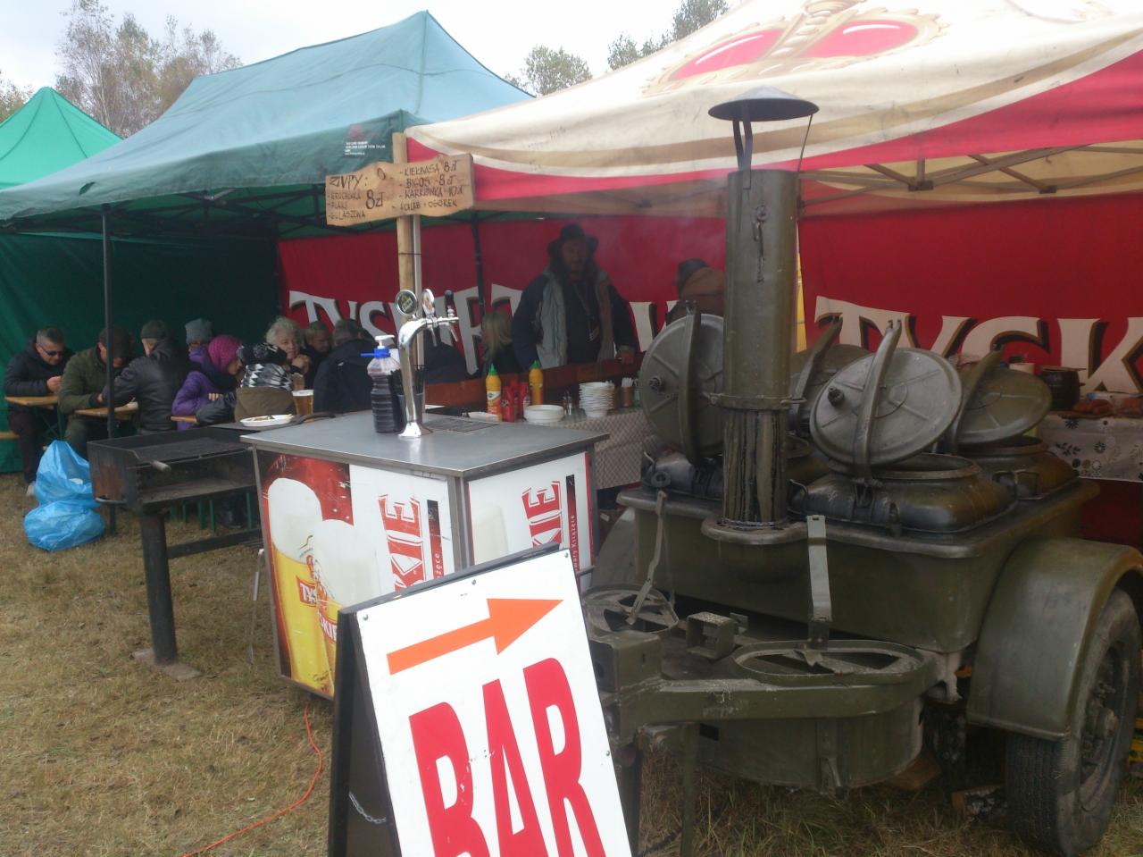 Polowa kuchnia wojskowa z parującymi kotłami, ustawiona obok baru z piwem i stolików pod namiotami podczas plenerowej imprezy.