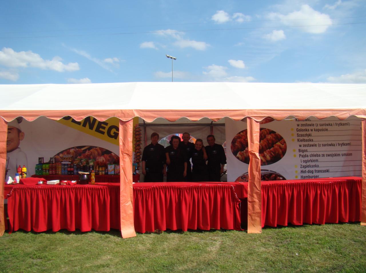 Ekipa gastronomiczna w czarnych uniformach za czerwonym stołem w namiocie z menu potraw na ścianie, widok z zewnątrz na trawnik i błękitne niebo.