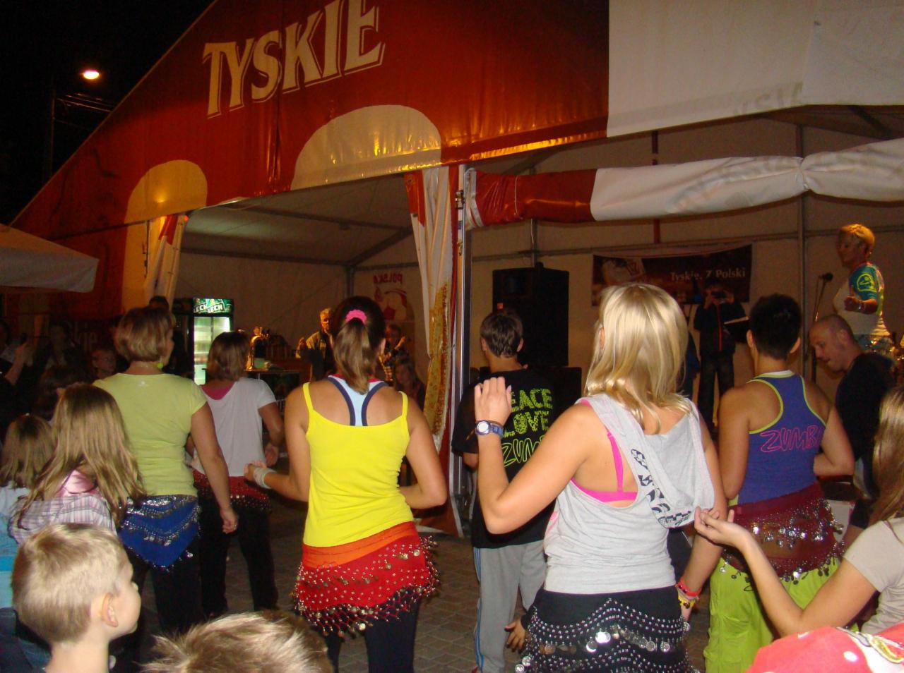 Grupa osób tańczących wewnątrz namiotu z logo Tyskie, widoczne kolorowe stroje taneczne z cekinami, scena z muzykami w tle.