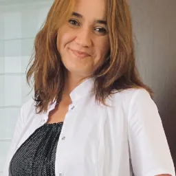 Uśmiechnięta kobieta w białym fartuchu medycznym, spiętym na guziki, z brązowymi włosami i czarną bluzką w białe kropki.