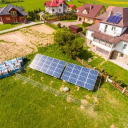 Widok z lotu ptaka na dwa panele fotowoltaiczne na zielonej łące, ogrodzone metalowym płotem, z widocznymi domami w tle i stertą materiałów budowlanych przykrytych plandeką obok paneli.