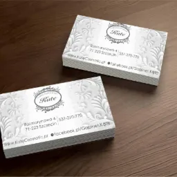 Dwa stosy wizytówek z wypukłym, białym wzorem kwiatowym i danymi kontaktowymi gabinetu kosmetycznego Kate na drewnianym tle.
