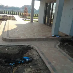 Ułożona kostka brukowa w odcieniach beżu i brązu tworzy ścieżkę i taras przy nowoczesnym domu. Widoczne przygotowania do systemu nawadniania w wykopie.