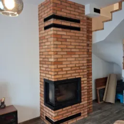 Murowany kominek z cegły klinkierowej z czarnymi wstawkami, widok z boku, na podłodze panele imitujące drewno, w tle schody i lampa sufitowa.