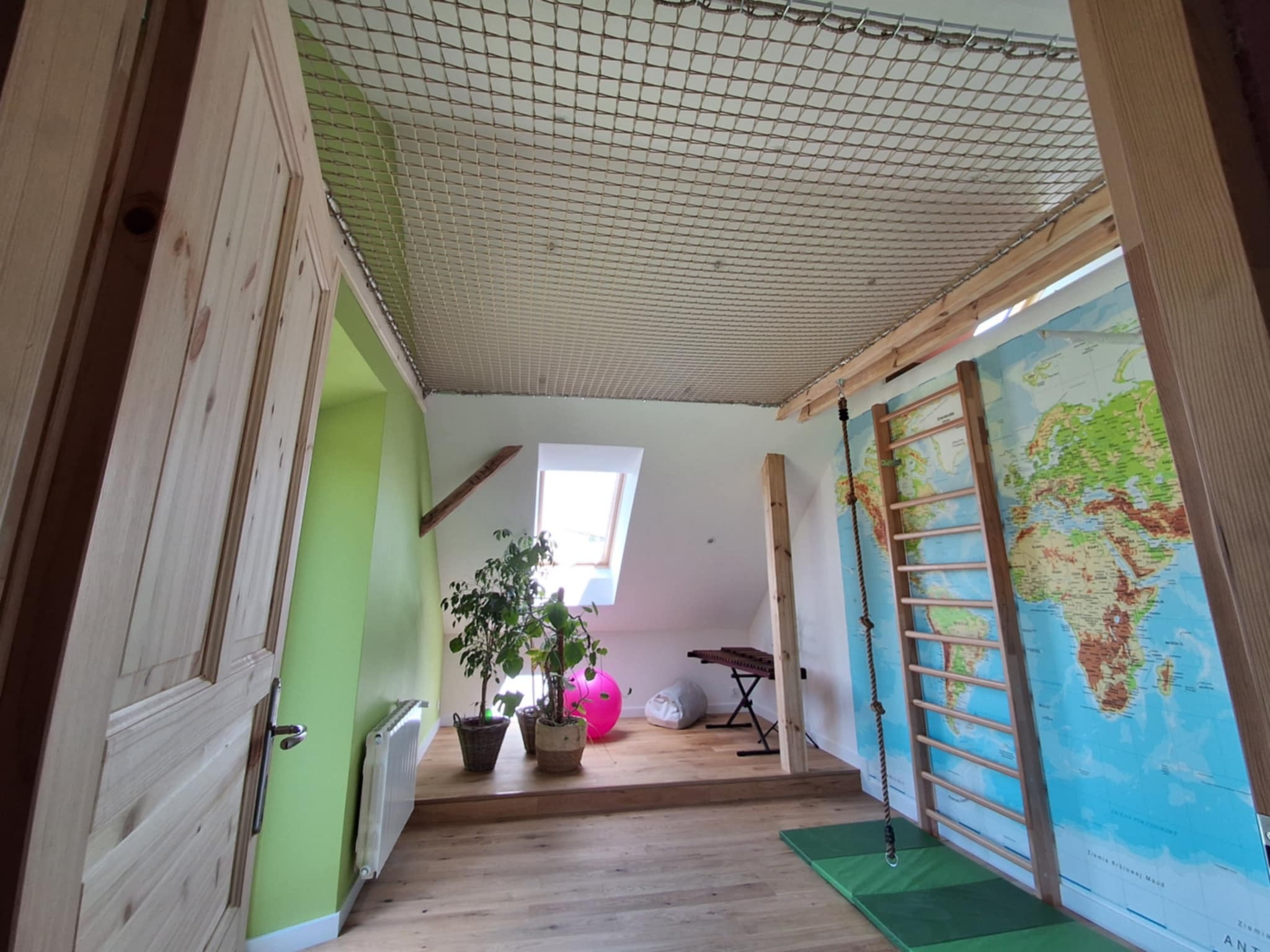 Pokój dziecięcy na poddaszu z siatką zabezpieczającą na suficie, drabinką gimnastyczną na ścianie z mapą świata i drewnianą podłogą.