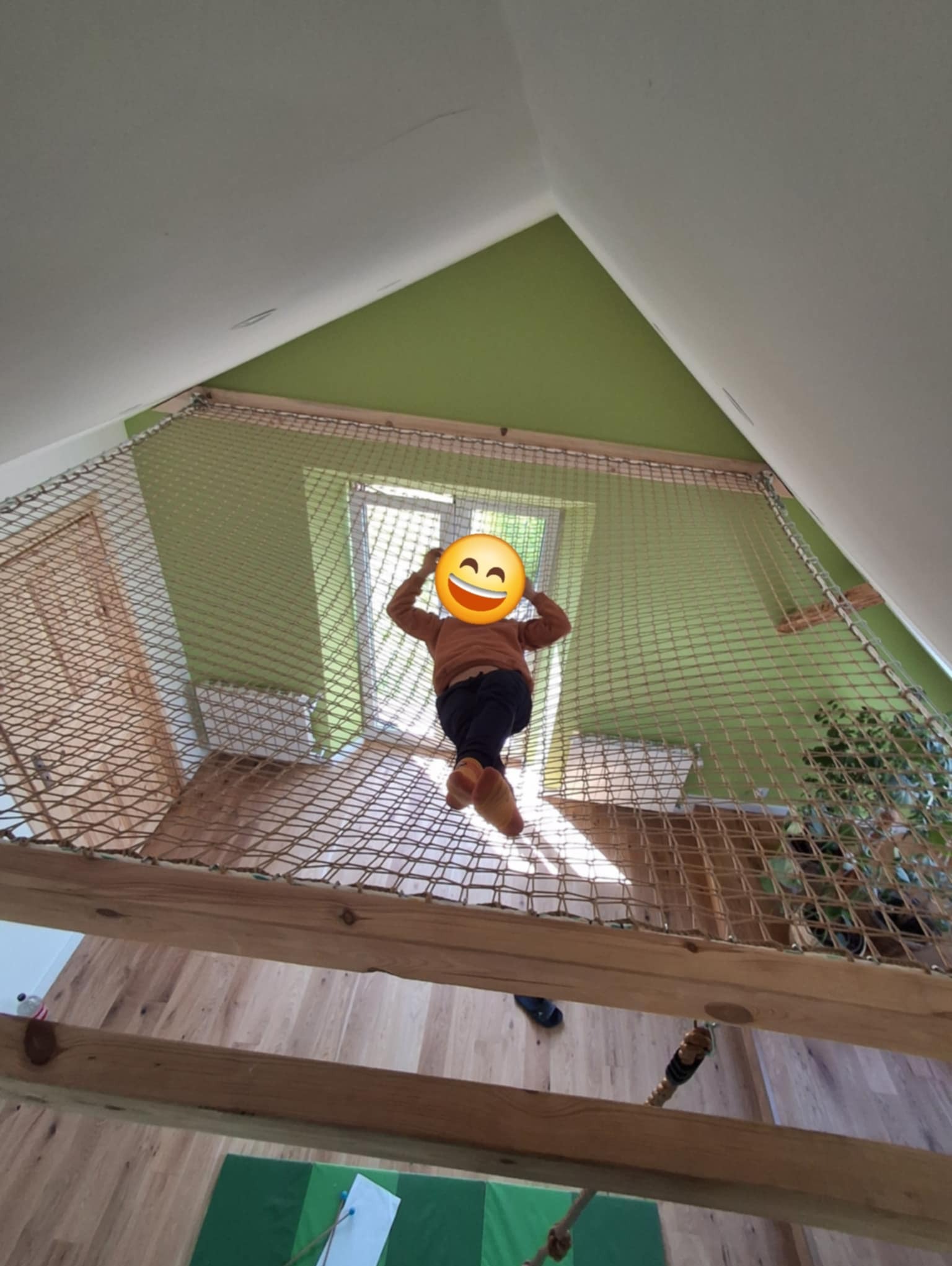 Pokój dziecięcy z siatką zabezpieczającą na antresoli, widok z dołu na drewnianą konstrukcję i zielone ściany. Nietypowy projekt wnętrza.