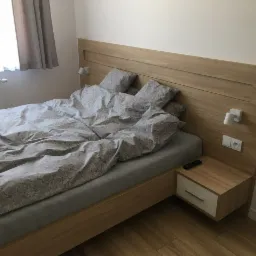 Drewniane łóżko z szarą pościelą w pokoju, zintegrowane szafki nocne z lampkami i gniazdkami.