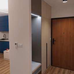 MEB-MAR - Nowoczesna szafa wnękowa z siedziskiem w przedpokoju. Minimalistyczny design, oświetlenie LED, drewniana podłoga i drzwi. Widoczny fragment niebieskiej kuchni.