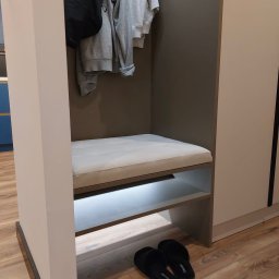 MEB-MAR - Szafa wnękowa z siedziskiem i oświetleniem LED, w kolorach szarości i bieli. Na półce pod siedziskiem leżą czarne kapcie. U góry wieszaki z ubraniami.