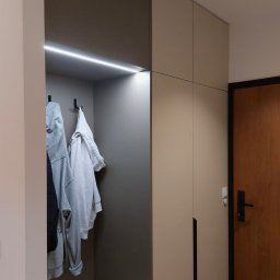 MEB-MAR - Nowoczesna szafa wnękowa z oświetleniem LED i wieszakami na ubrania. Minimalistyczny design, jasne kolory, widoczna część drzwi wejściowych.