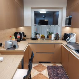 MEB-MAR - Nowoczesna kuchnia w kształcie litery U z beżowymi frontami, drewnianym blatem i oświetleniem LED. Na blacie ekspres do kawy i inne akcesoria. Na podłodze dywanik z motywem kawy.