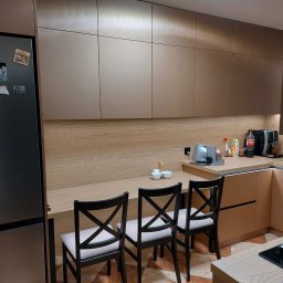 MEB-MAR - Nowoczesna kuchnia na wymiar z zabudową w odcieniach beżu i drewna, zintegrowana z blatem pełniącym funkcję barku i trzema krzesłami. Widoczny sprzęt AGD i akcesoria.