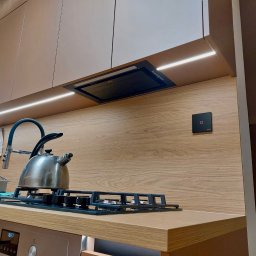 MEB-MAR - Nowoczesna kuchnia z drewnianym blatem i oświetleniem LED w Radomsku. Na blacie czajnik i płyta gazowa, nad nimi okap. Jasne fronty szafek górnych.