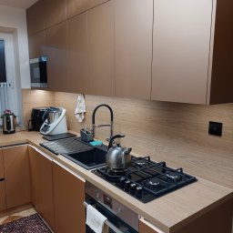 MEB-MAR - Nowoczesna kuchnia z zabudową w odcieniach beżu i drewna. Górne szafki bez uchwytów, czarny zlew i płyta gazowa. Na blacie czajnik i akcesoria kuchenne.