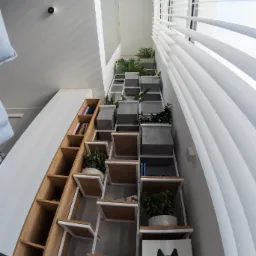 Wielofunkcyjne schody z drewnianymi półkami i białym metalowym stelażem, wykorzystane jako regał na książki, doniczki z roślinami i legowisko dla kota, widok z dołu w kierunku okna z żaluzjami.