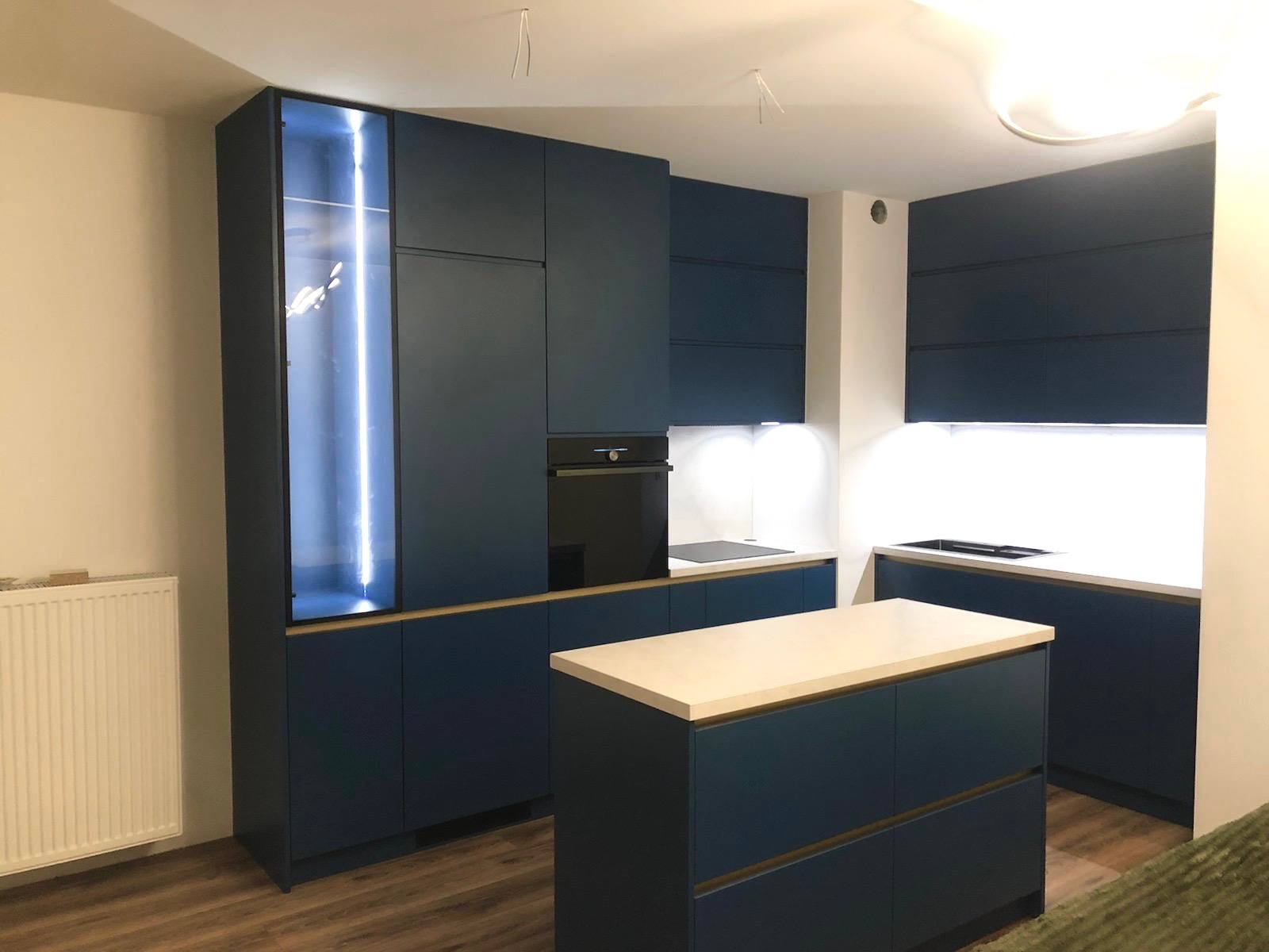 Granatowa kuchnia z wyspą i podświetlaną szafką. Nowoczesne meble kuchenne z jasnym blatem i drewnianymi akcentami. Minimalistyczny design, oświetlenie LED.