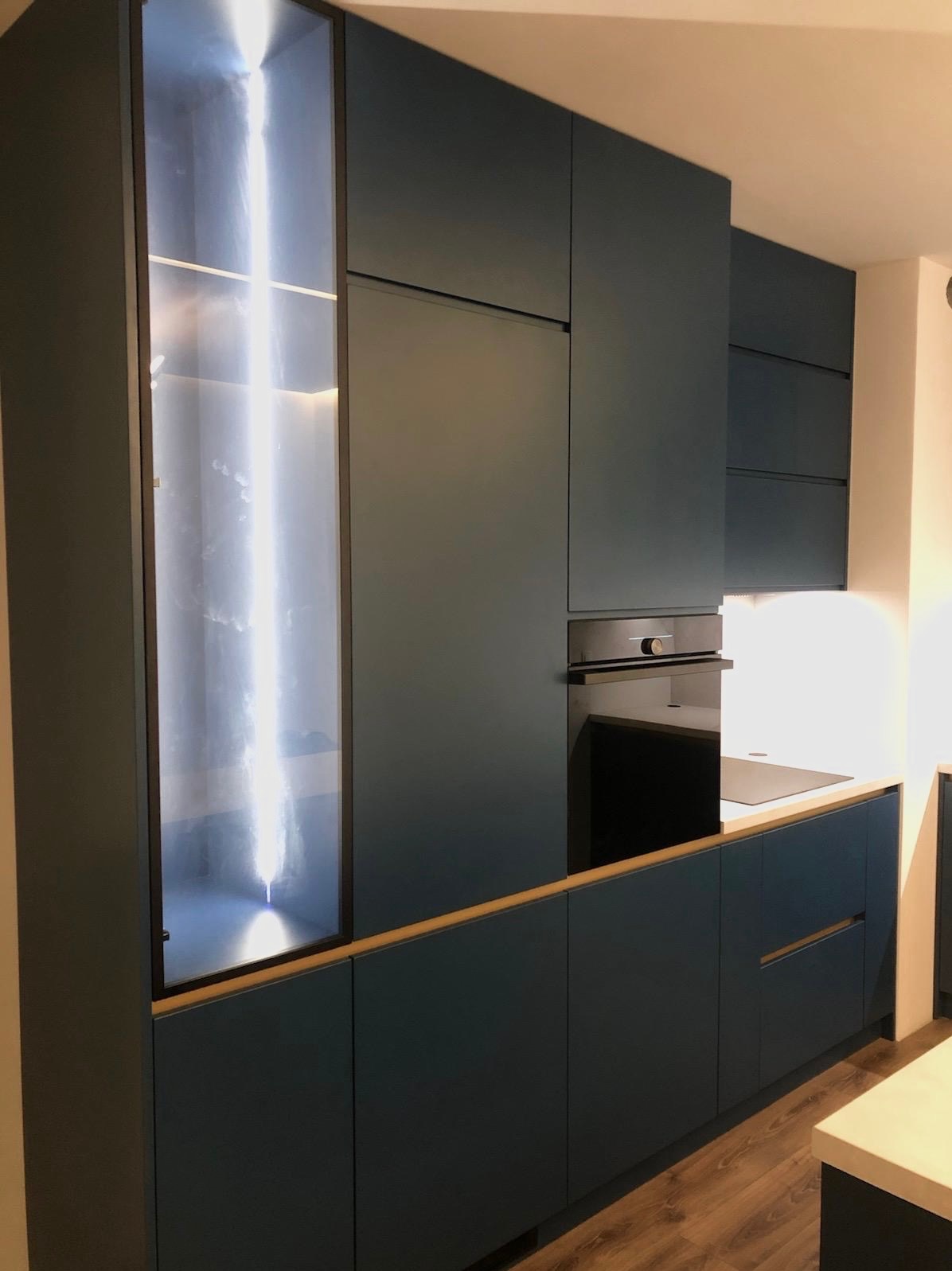 Nowoczesna, granatowa zabudowa kuchenna z podświetlaną witryną i drewnianym blatem. Minimalistyczny design, piekarnik w zabudowie, drewniana podłoga.