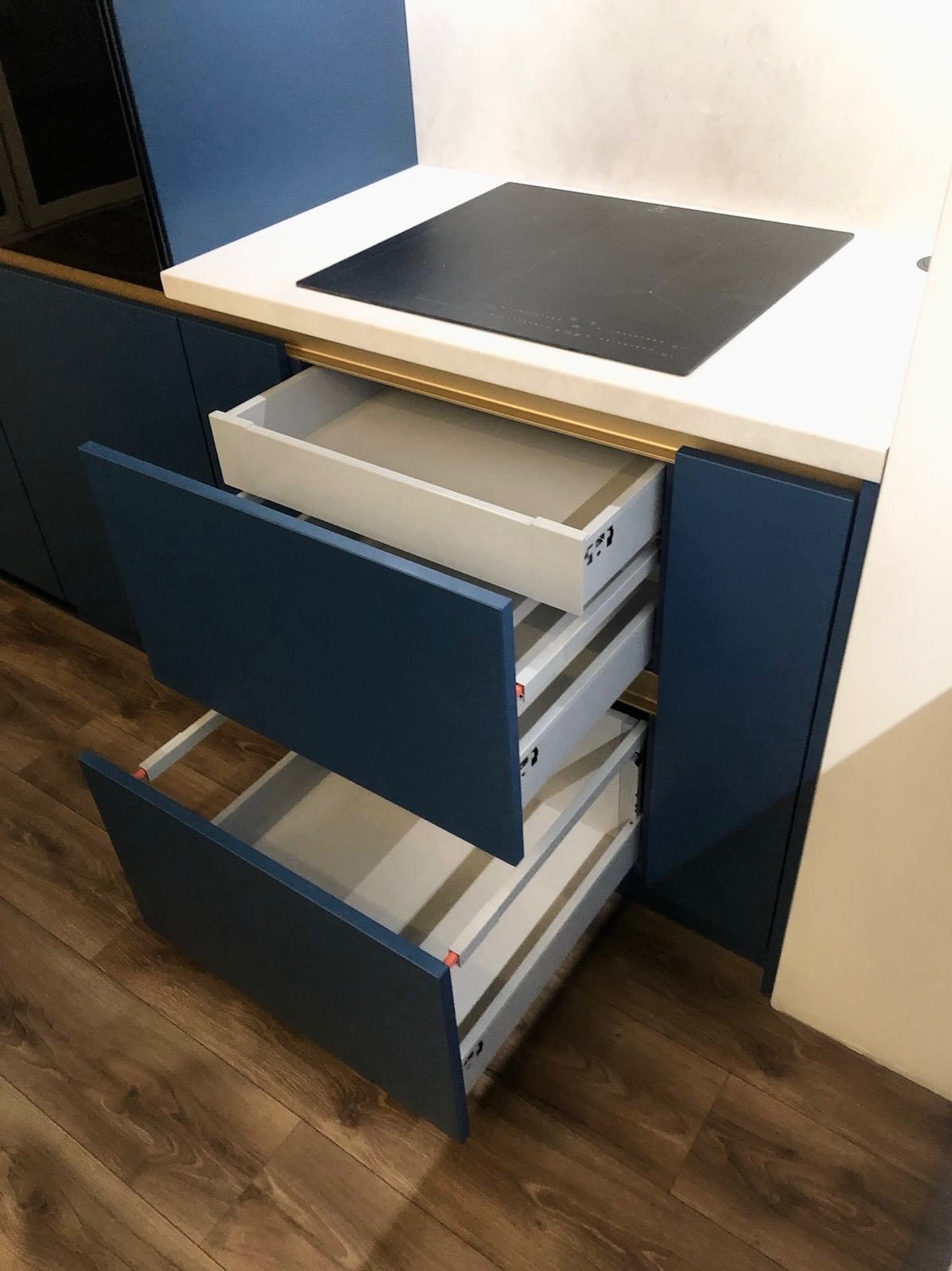 Niebieska zabudowa kuchenna z otwartymi szufladami, widoczna płyta indukcyjna na blacie. Szuflady z białym wnętrzem. Ujęcie z bliska, ukazujące detale wykończenia.