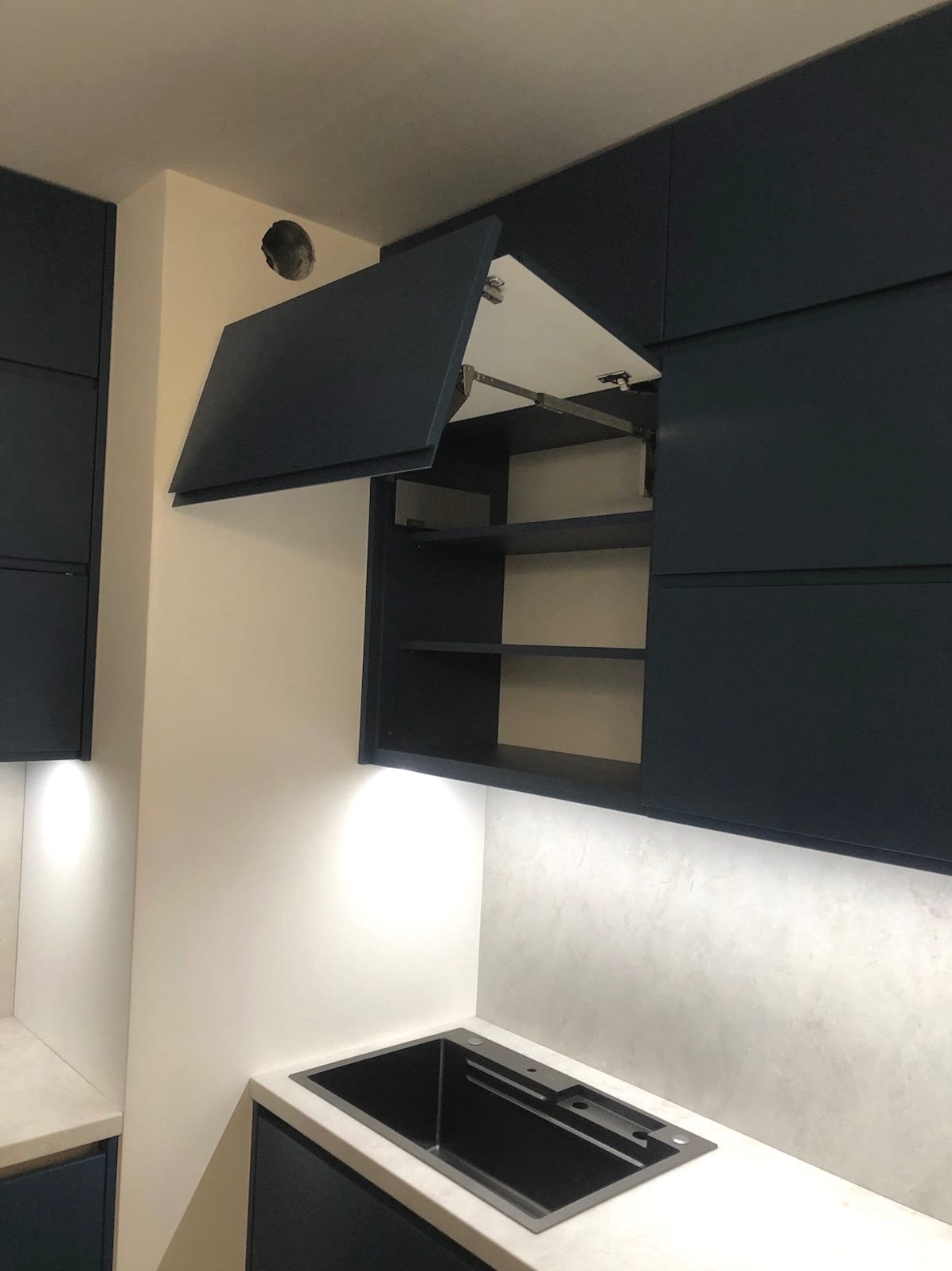 Granatowa zabudowa kuchenna z otwartą szafką górną, widoczne półki i mechanizm podnoszenia frontu. Podświetlenie LED pod szafkami, zlew w blacie.