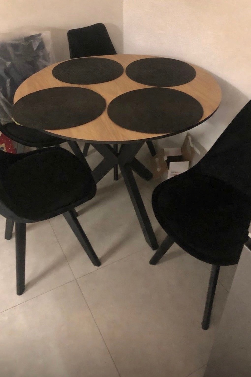 Okrągły stół z jasnym blatem i czarną, geometryczną podstawą, otoczony czarnymi krzesłami z welurowym obiciem i okrągłymi podkładkami. Minimalistyczny design.