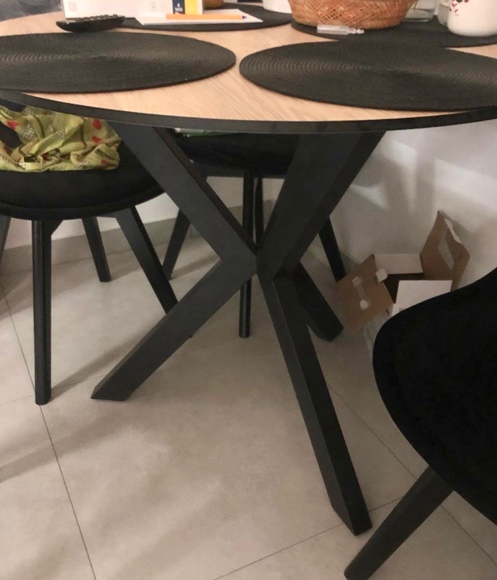 Okrągły stół z jasnym blatem i czarnymi, krzyżującymi się nogami, otoczony czarnymi krzesłami. Widoczne nakrycia i elementy dekoracyjne na blacie.