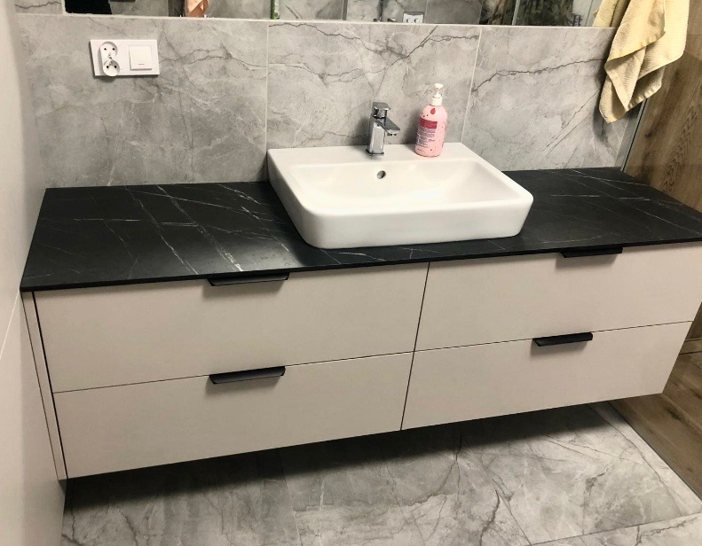 Biała umywalka nablatowa na szafce z czterema szufladami i czarnym blatem imitującym marmur. W tle szare płytki z marmurowym wzorem i fragment lustra.