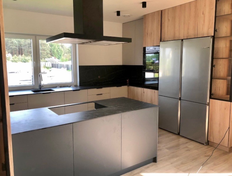 Nowoczesna kuchnia z wyspą, fronty w odcieniach szarości i drewna, czarny backsplash i okap. Duża lodówka side-by-side. Naturalne światło z okna.