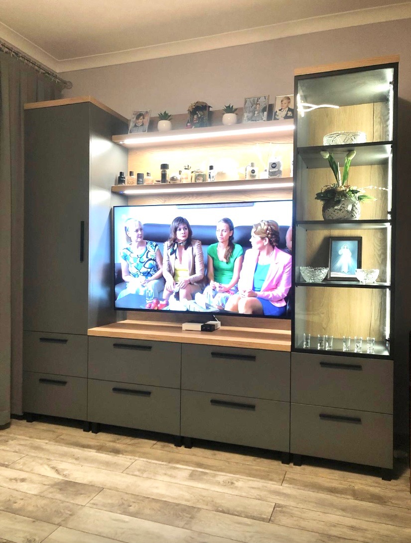 Szara zabudowa meblowa z drewnianymi akcentami, oświetleniem LED i szklaną witryną, z telewizorem wkomponowanym w centralną część. Nowoczesny design.