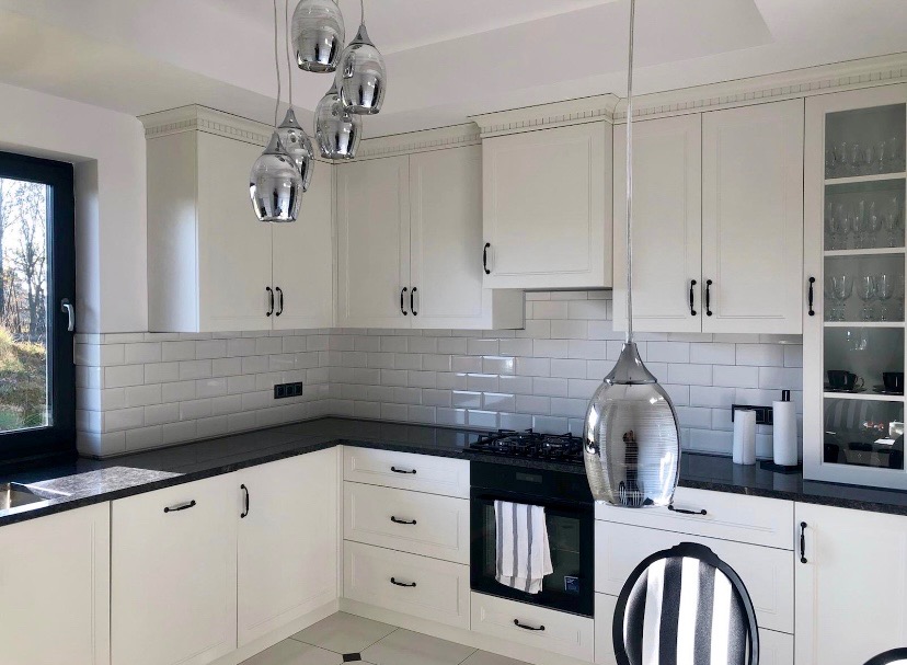 Biała kuchnia z czarnymi uchwytami i blatem, oświetlona wiszącymi lampami. Widoczne płytki na ścianie i fragment krzesła. Nowoczesny design.