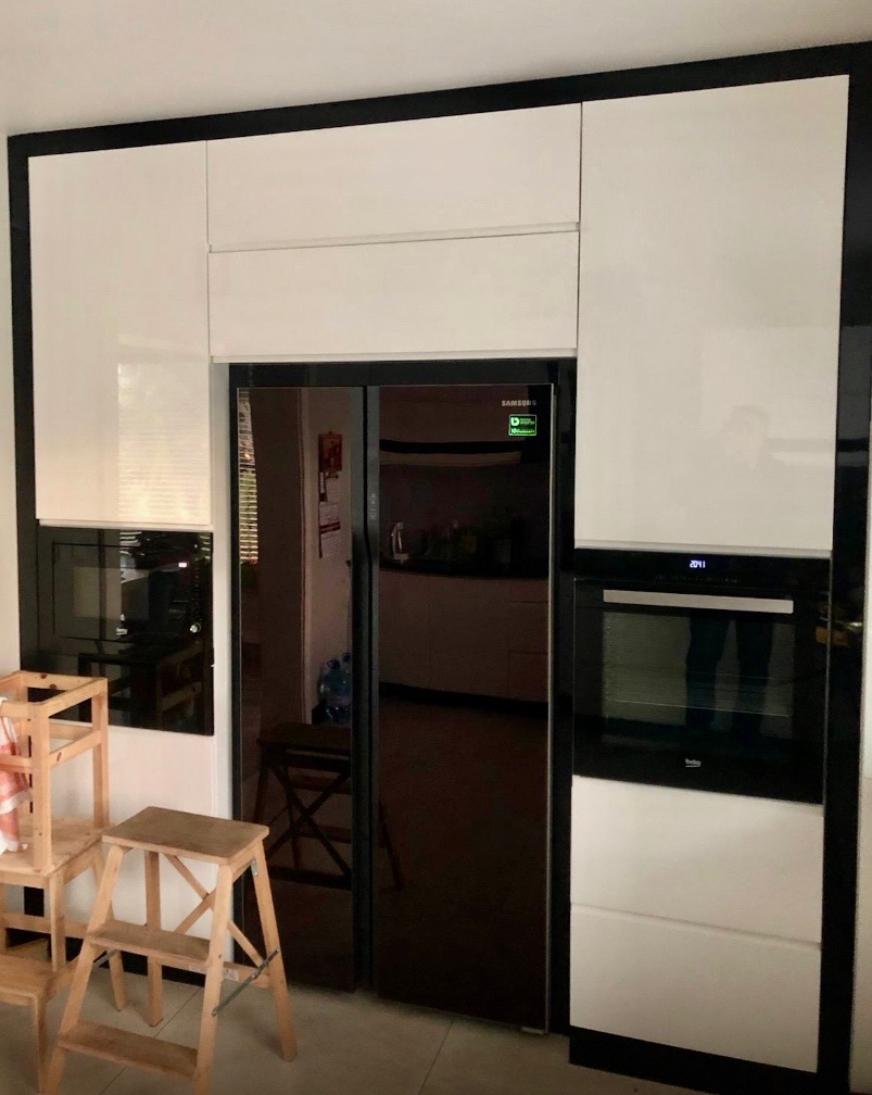 Biała zabudowa kuchenna z czarnymi obramowaniami, wkomponowana lodówka i piekarnik. W tle widoczne schodki.