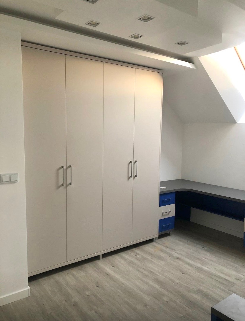 Biała szafa wnękowa z czterema drzwiami i srebrnymi uchwytami, dopasowana do skosu dachu, z biurkiem w kolorze szaro-niebieskim w tle.
