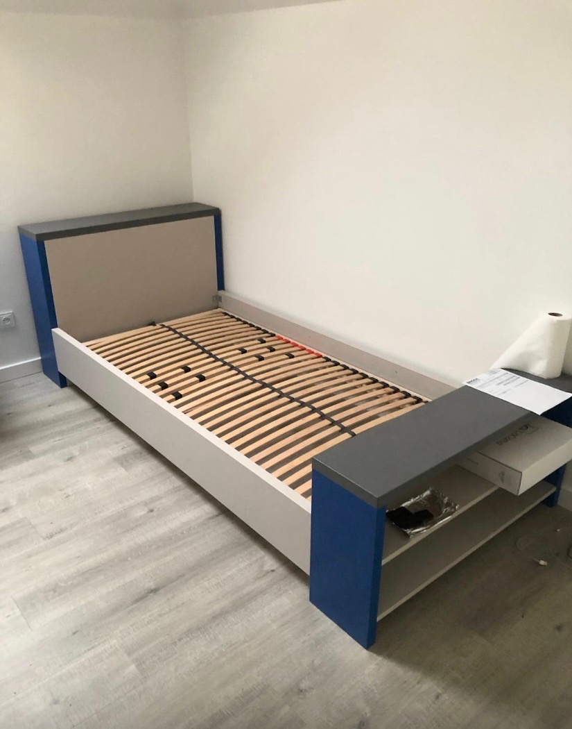 Niebiesko-szare łóżko jednoosobowe z drewnianym stelażem i półką nocną, ustawione na panelach podłogowych w jasnym pokoju. Minimalistyczny design.