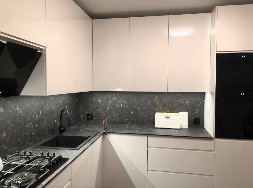 Biała, nowoczesna kuchnia narożna z czarnym blatem imitującym marmur i czarną armaturą. Widoczny fragment płyty gazowej i piekarnik.