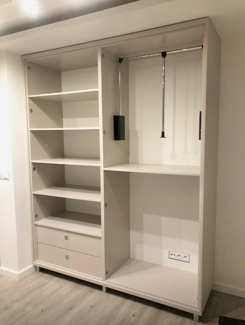 Jasna szafa na wymiar z otwartymi drzwiami, widoczne półki, szuflady i drążek na ubrania. Minimalistyczny design, gładkie fronty, subtelne nóżki. W tle ściana i gniazdko elektryczne.