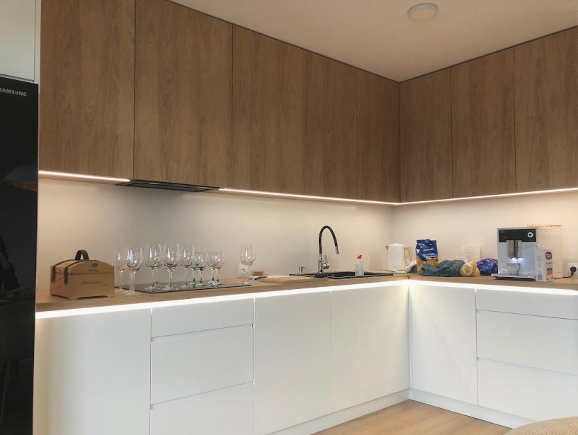 Nowoczesna kuchnia narożna z białymi frontami i drewnianymi szafkami górnymi, podświetlona listwami LED. Na blacie szklanki i akcesoria kuchenne.