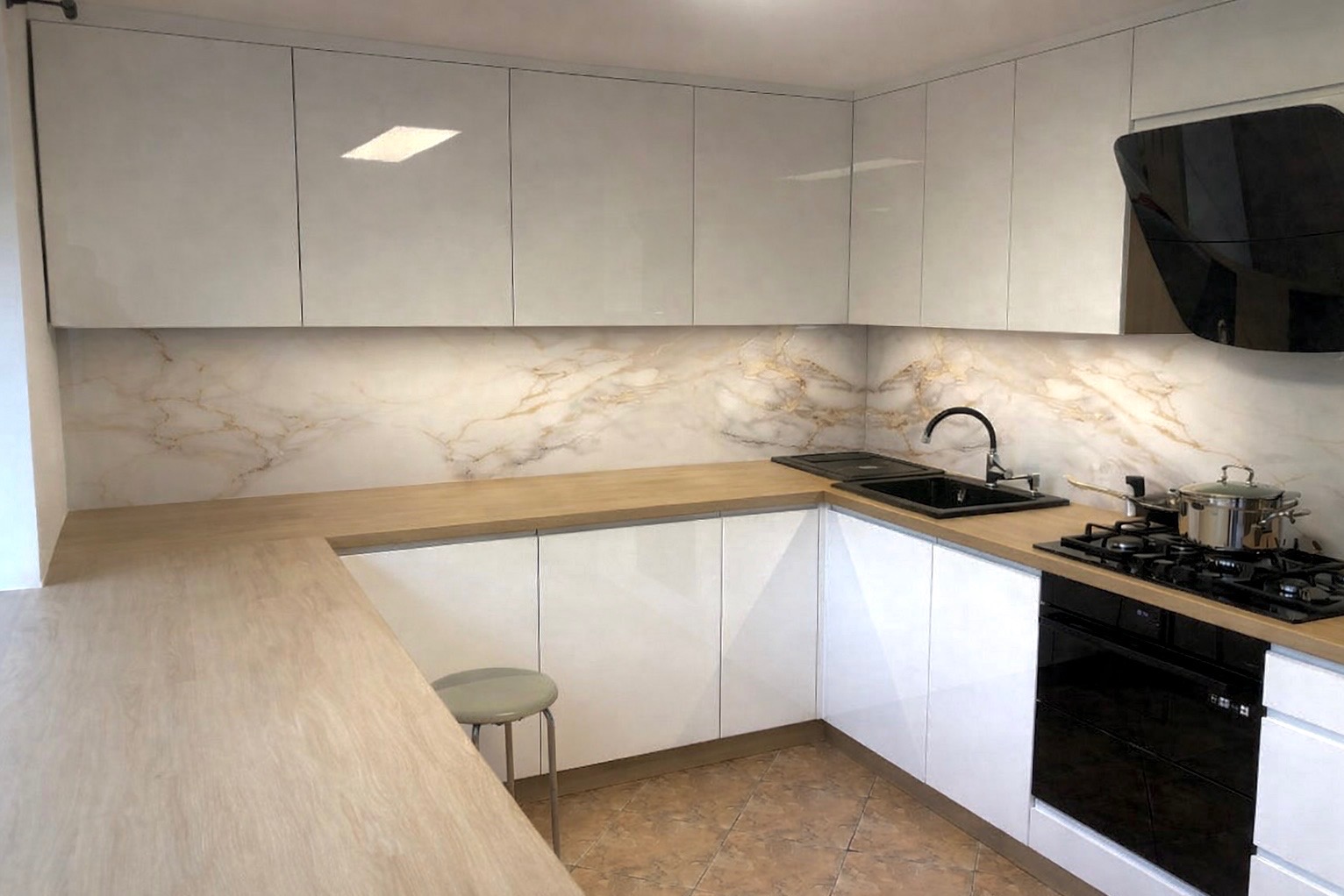 Biała kuchnia z połyskiem, drewniany blat i marmurowy backsplash. Na blacie czarny zlew i płyta grzewcza z garnkiem. W tle stołek barowy.