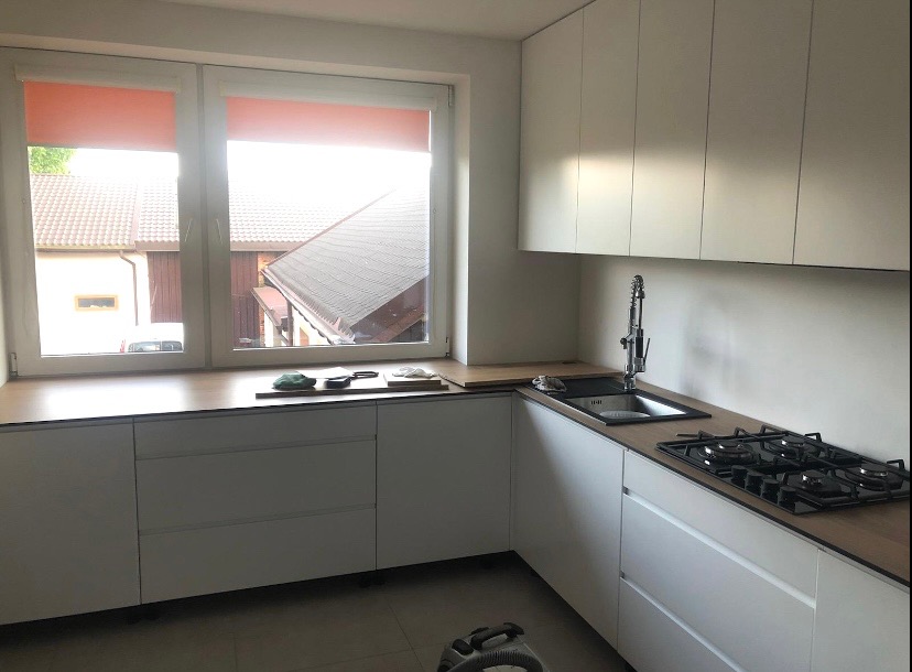 Jasna kuchnia z białymi frontami i drewnianym blatem, widok na okno z roletami. Minimalistyczny design, nowoczesne wyposażenie, czarny zlew i płyta gazowa.