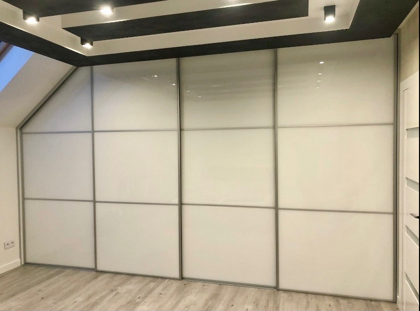 Biała szafa wnękowa z przesuwnymi drzwiami, dopasowana do skosu poddasza. Metalowe ramy drzwi, minimalistyczny design, oświetlenie sufitowe.