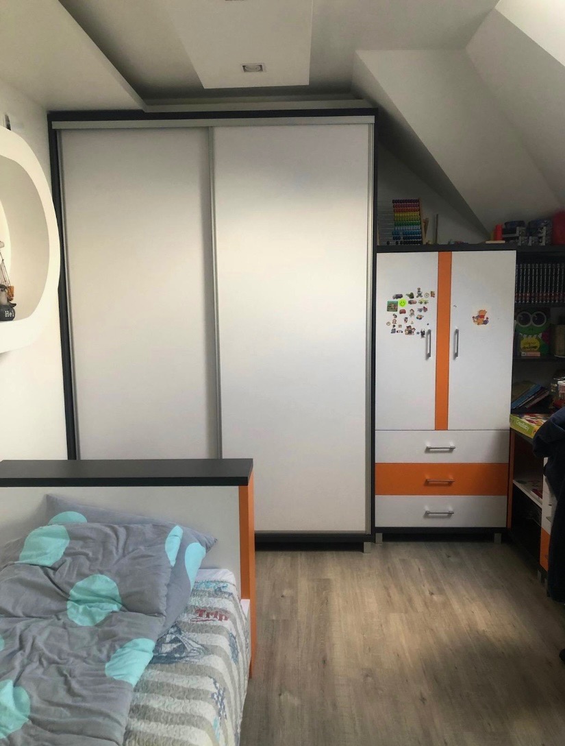 Biała szafa przesuwna z czarnymi obramowaniami w pokoju na poddaszu, obok łóżko z szarą pościelą w kropki i biała szafka z pomarańczowymi akcentami.