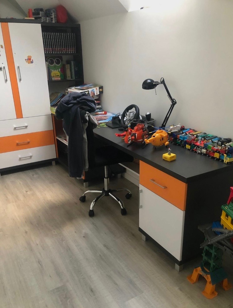 Pokój dziecięcy z biurkiem i szafą w kolorach bieli i pomarańczy. Na biurku zabawki, lampka i kierownica do gier. Podłoga wyłożona panelami w odcieniach szarości.