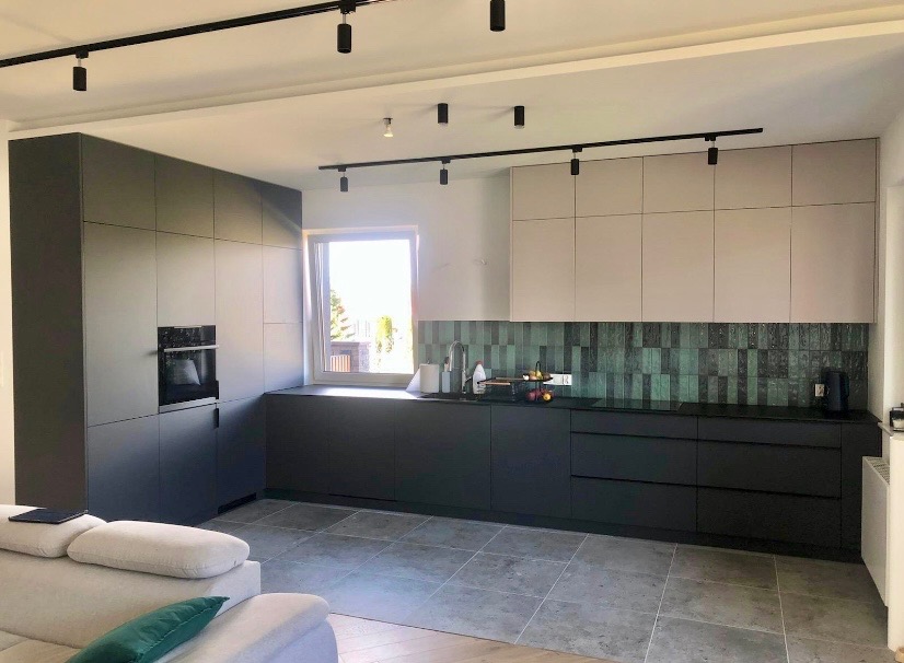 Nowoczesna kuchnia z szarymi szafkami, czarnym blatem i zieloną mozaiką na ścianie. Oświetlenie punktowe na suficie. Widok na okno i fragment sofy.