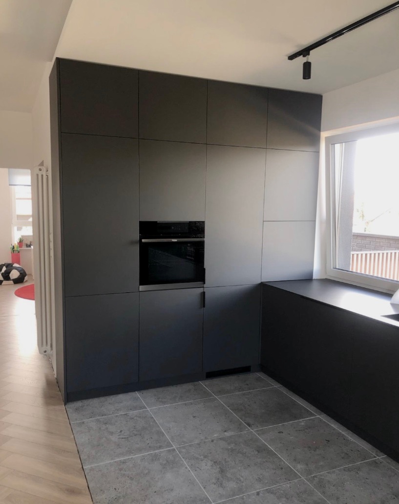 Grafitowa zabudowa kuchenna z piekarnikiem w nowoczesnym wnętrzu. Minimalistyczny design, szare płytki podłogowe, światło dzienne z okna.
